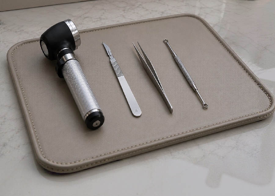 A dermatoscope, scalpel, forceps, and curette arranged on a soft leather tray